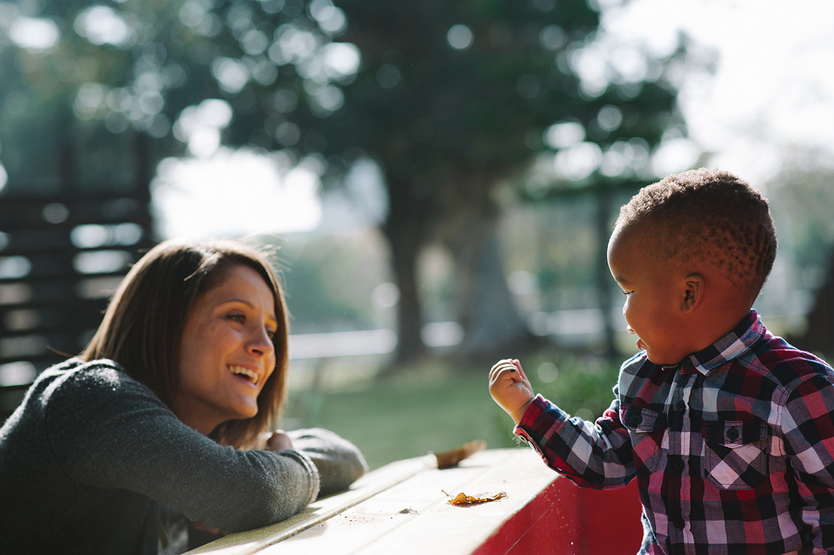 A smiling woman with arms folded on an outdoor table looks at a young boy