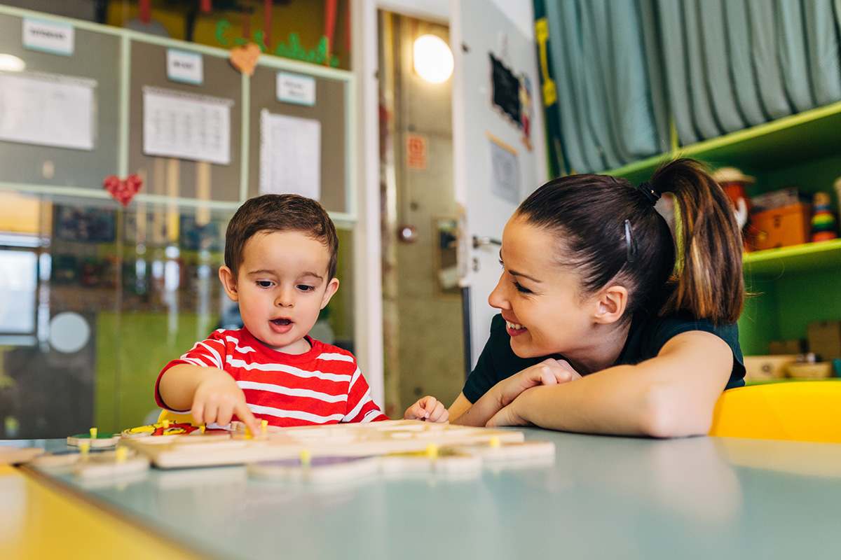 A young boy plays at a table in an early care center as a smiling teacher looks on