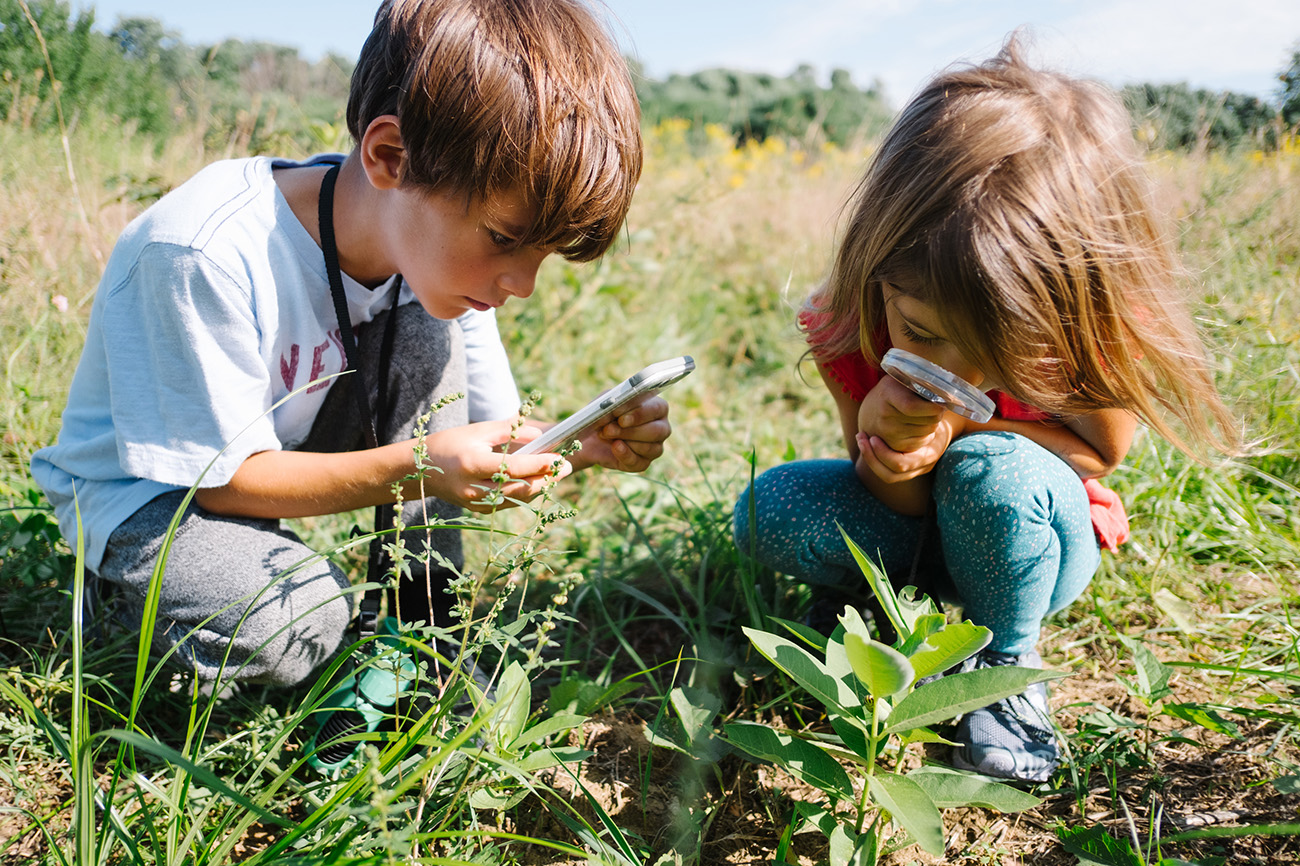 A preschool-age boy and girl squat in the tall grass, each looking at plants through magnifying glasses