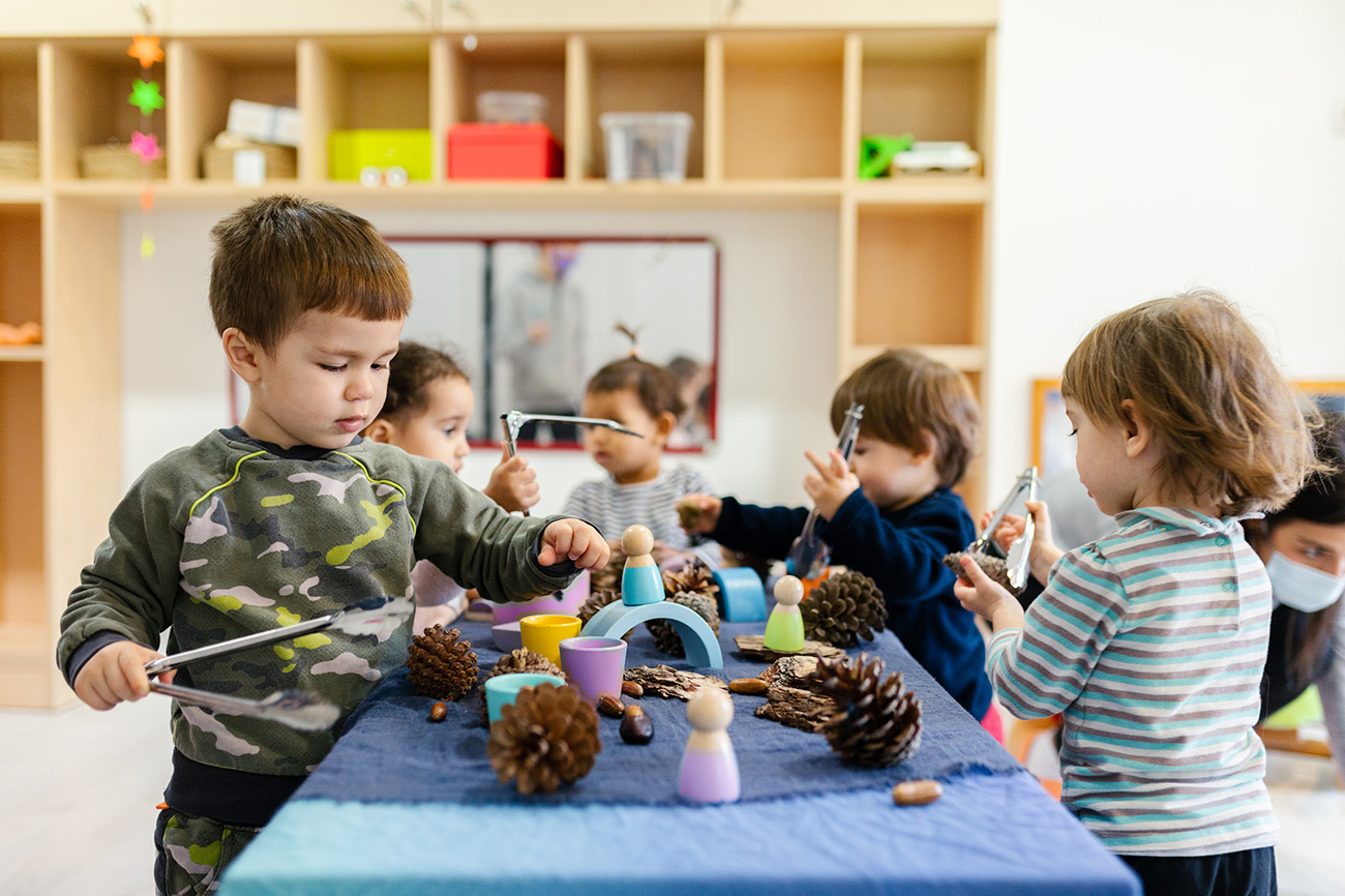 Preschool-age children play at an indoor table at an early care center, examining pine cones with tongs