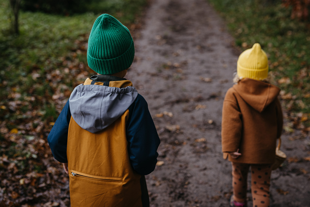 A preschool-age boy and girl wearing coats and hats walk away from the camera, down a dirt path in the fall
