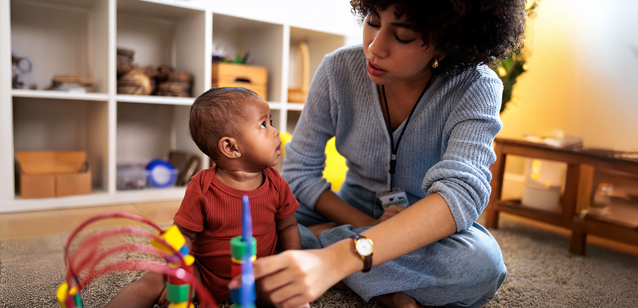 A caregiver is helping a baby learn to stack colorful rings on a toy. The caregiver is sitting cross-legged on the floor, demonstrating how to place the rings on the vertical post, while the baby watches intently, absorbing the new skill.